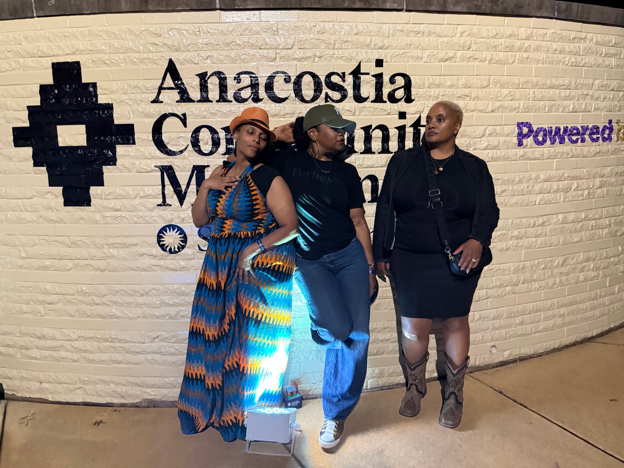 From left, Dr. Tonie Tyler, Precious “Preciosa” Myers-Brown, and KV Simmons pose at night in front of the Smithsonian’s Anacostia Community Museum sign on a cream brick wall. Dr. Tonie Tyler wears a blue and orange print maxi dress with an orange hat; Precious wears a black tee, jeans, and a cap; KV wears all black with cowboy boots.