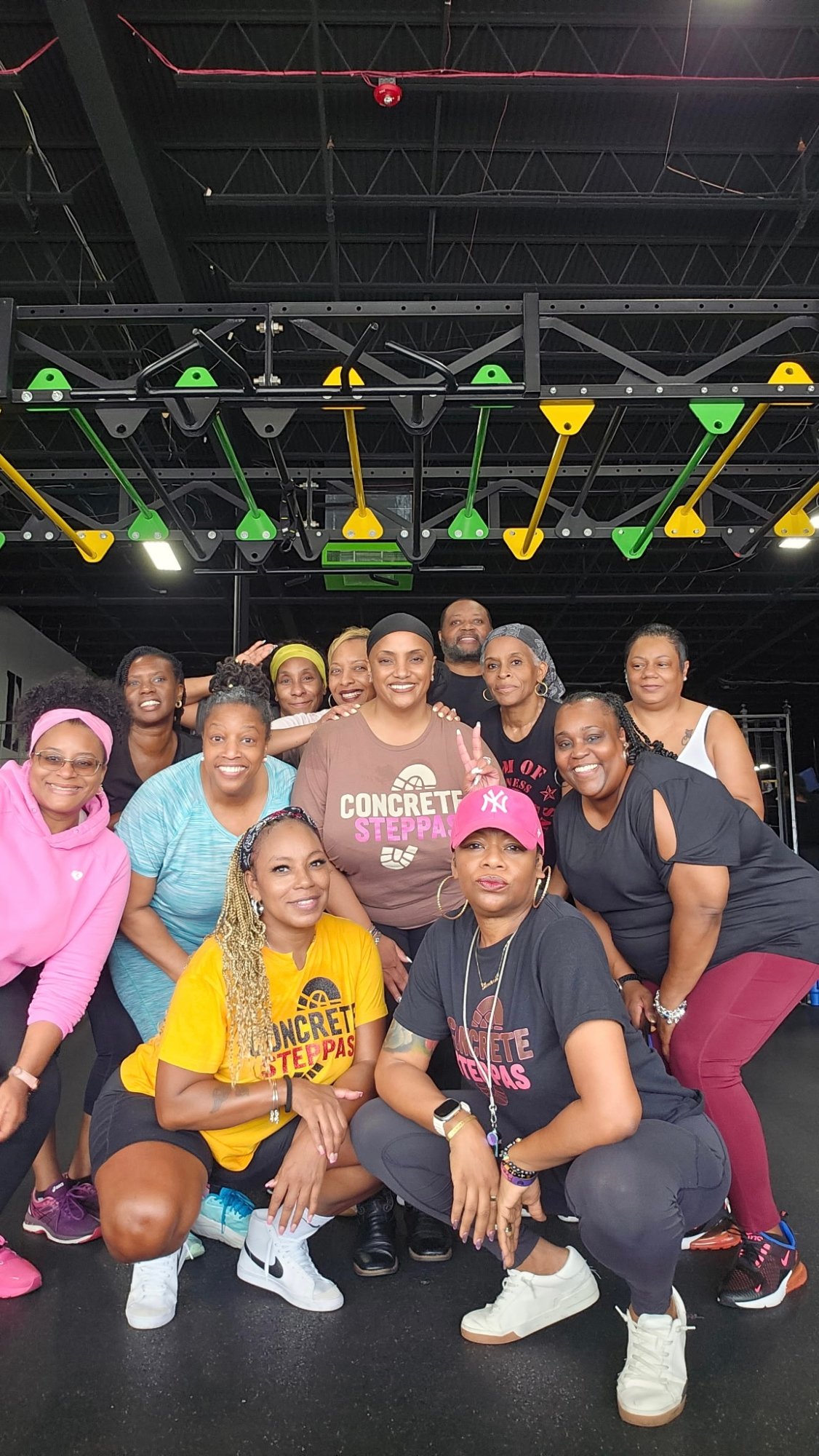 A group poses together in a gym beneath exercise equipment. Several people wear Concrete Steppas™ shirts, with others in colorful workout clothes.